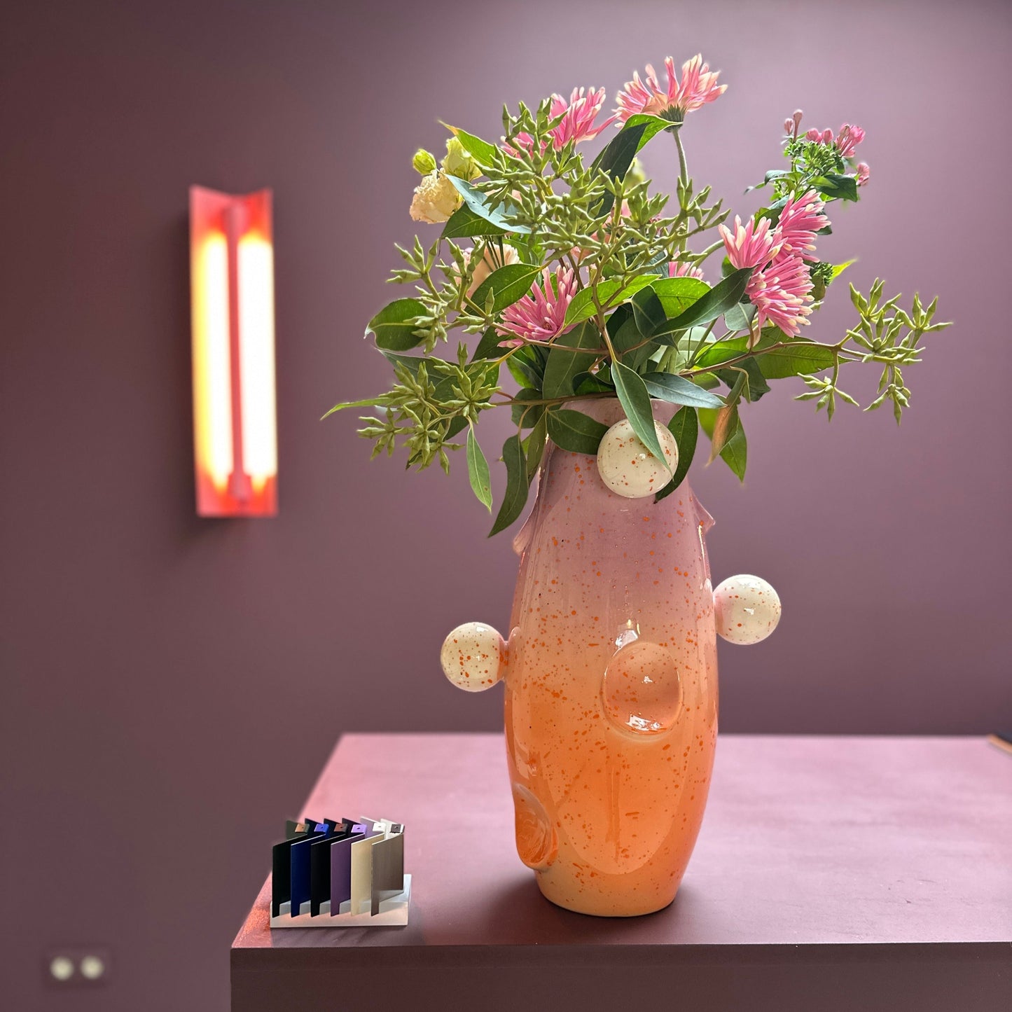 RUIMTE — OKO Big Circus vase by Malwina Konopacka, a unique ceramic piece with oval recesses, gradient orange-lilac tones, and multicolored ball motifs, styled on a wooden table with a contemporary pink lamp in the background.
