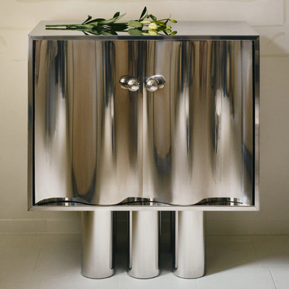 MOTION commode in polished stainless steel with curved wave-front doors, round knobs and cylindrical legs, styled with flowers, by The Good Living & Co.