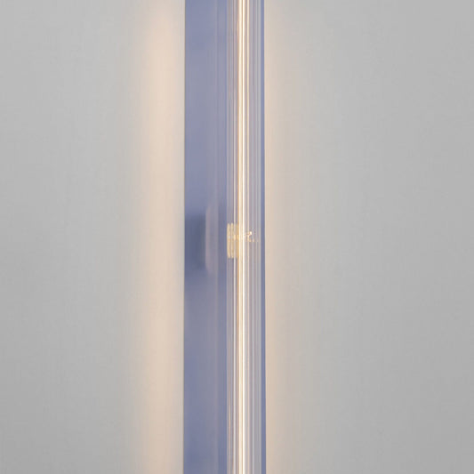 A modern vertical wall sconce with a translucent lavender-blue rectangular housing and a glowing warm white tube light inside, mounted on a plain light gray wall.