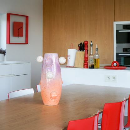 RUIMTE — OKO Big Circus vase by Malwina Konopacka, a unique ceramic piece with oval recesses, gradient orange-lilac tones, and multicolored ball motifs, styled on a wooden table in a kitchen with red chairs and a counter in the background.
