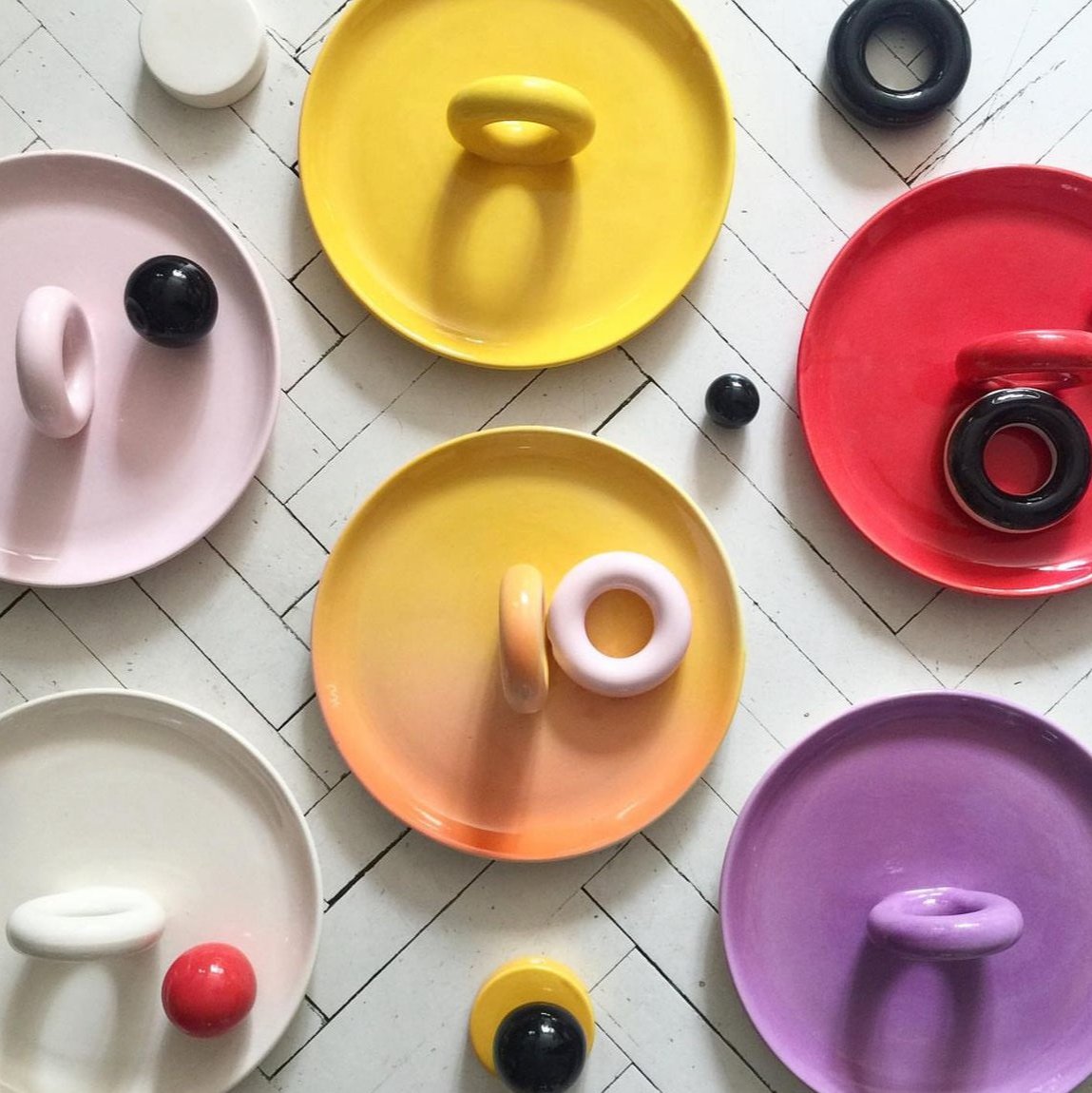 A collection of various colored ceramic fruit plates by Malwina Konopacka with round handles and unglazed rims, displayed on a white grid background.