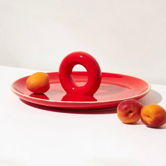 Red ceramic fruit platter with a round handle, featuring two peaches on top, photographed against a clean white background — as featured in Knack Weekend.